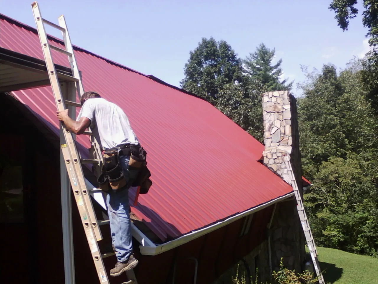 Roofer on a ladder beside a red metal roof and stone chimney — Soffit & Fascia Repair by Roofing Brothers in Raeford